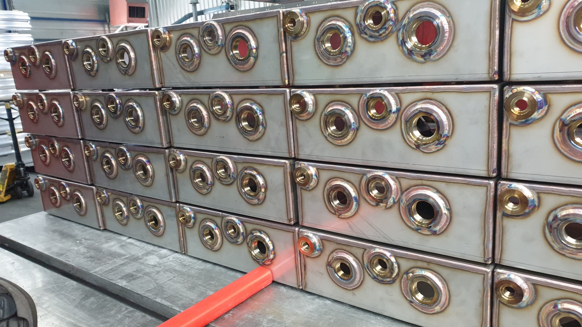 Równy rząd pospawanych, metalowych skrzynek z otworami o różnych średnicach, widoczne ślady spawania na obrzeżach otworów, całość ułożona na metalowym stole warsztatowym.