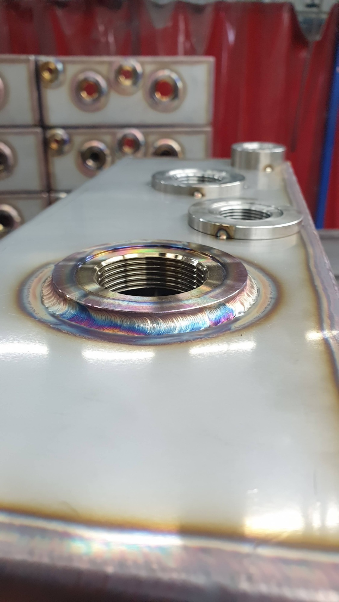 Zbliżenie na spawany gwintowany otwór w metalowym zbiorniku, z widocznymi tęczowymi przebarwieniami spoiny i precyzyjnymi ściegami spawalniczymi.