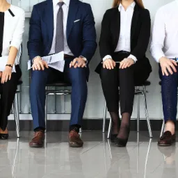 Czterech kandydatów siedzi na krzesłach, czekając na rozmowę kwalifikacyjną. Widoczne nogi i fragmenty ubrań, trzymają dokumenty i okulary.