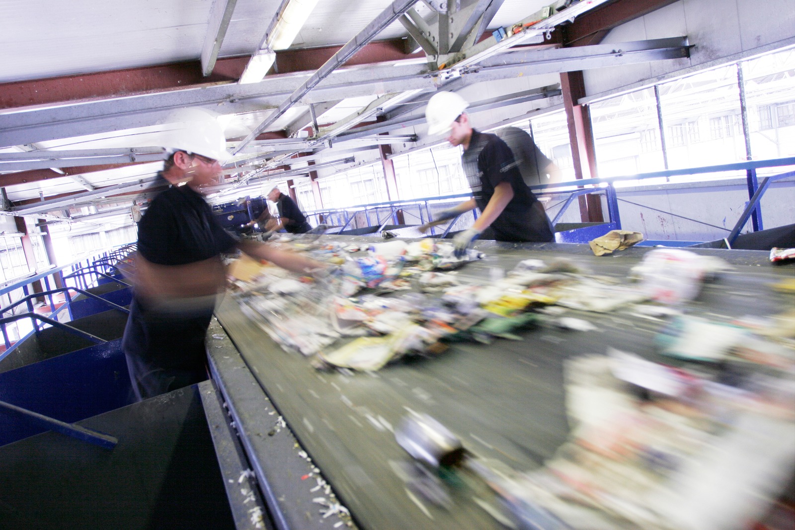 Pracownicy w białych kaskach sortują śmieci na taśmie produkcyjnej, w tle widoczne okna i konstrukcja hali.
