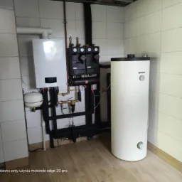 Kotłownia z białym kotłem gazowym, zasobnikiem ciepłej wody i czarną armaturą na tle białych płytek i drewnopodobnej podłogi.