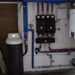 Refit Dawid Staszewski - Zestaw dwóch czarnych rozdzielaczy z manometrami i armaturą, podłączonych rurami w izolacji, obok zmiękczacz wody w piwnicy z płytkami podłogowymi.