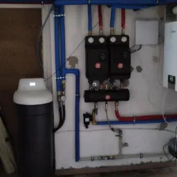 Zestaw dwóch czarnych rozdzielaczy z manometrami i armaturą, podłączonych rurami w izolacji, obok zmiękczacz wody w piwnicy z płytkami podłogowymi.