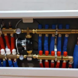 Rozdzielacz ogrzewania podłogowego w szafce, rury czerwone i niebieskie, manometry i pompa obiegowa. Widoczne białe głowice termostatyczne.