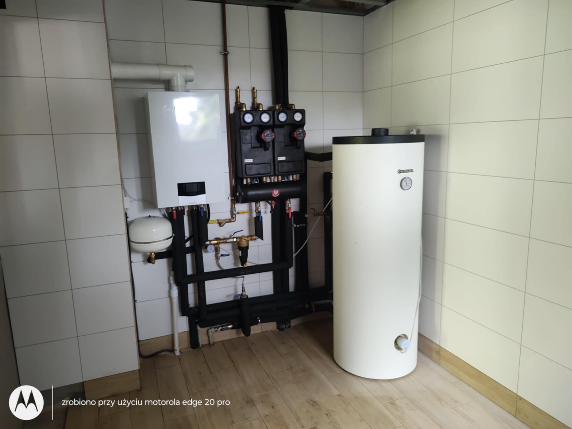Kotłownia z białym kotłem gazowym, zasobnikiem ciepłej wody i czarną armaturą na tle białych płytek i drewnopodobnej podłogi.