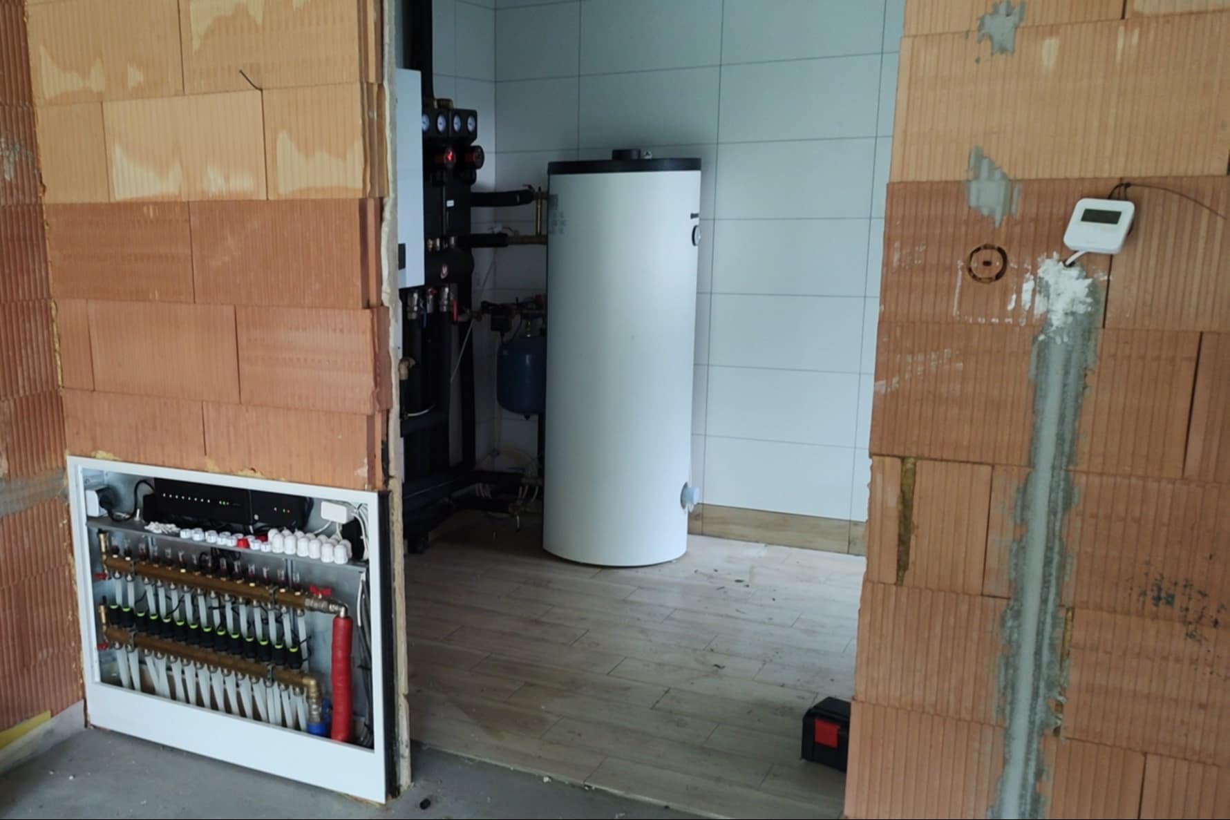 Kotłownia w trakcie budowy: widoczny biały zasobnik ciepłej wody, rozdzielacz ogrzewania podłogowego w ścianie z pustaków ceramicznych, czarna armatura i białe płytki na ścianie w tle.