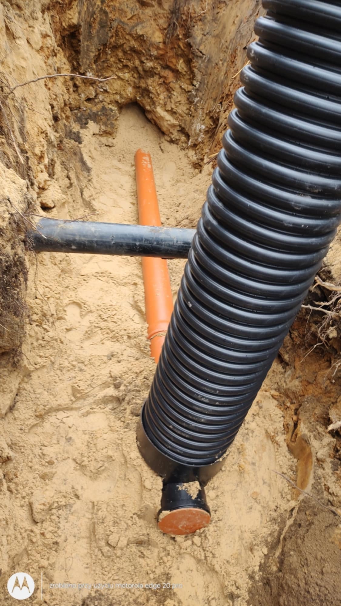 Wykop z widocznymi rurami: czarna karbowana rura kanalizacyjna pionowo, pomarańczowa rura pionowo w tle i czarna rura poziomo, całość w piaszczystym wykopie.