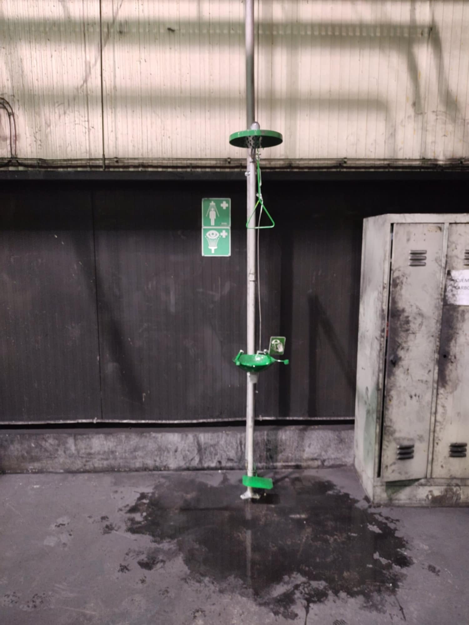 Awaryjny prysznic i płuczka do oczu w hali przemysłowej, widoczna kałuża wody na posadzce, obok metalowa szafka ubraniowa.
