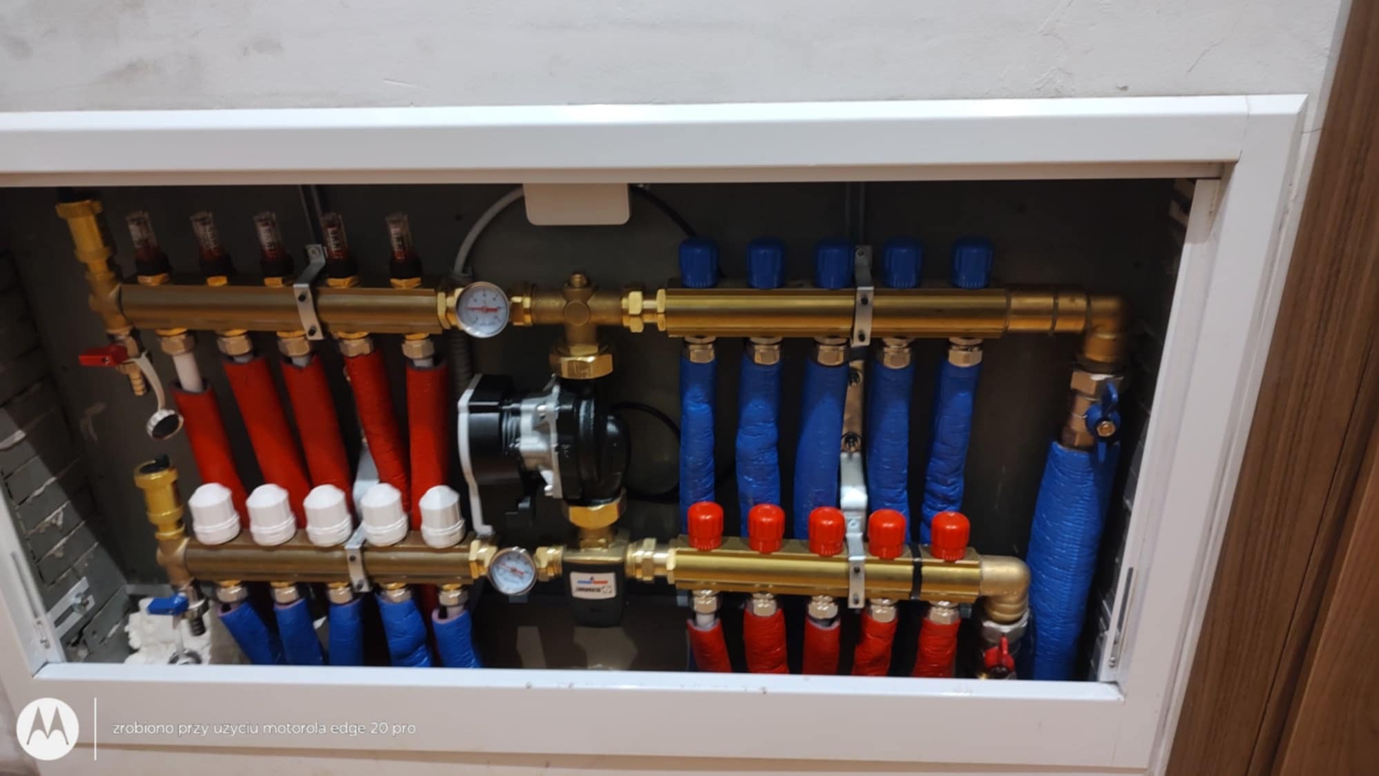 Rozdzielacz ogrzewania podłogowego w szafce, rury czerwone i niebieskie, manometry i pompa obiegowa. Widoczne białe głowice termostatyczne.