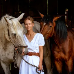 Portret kobiety w białej sukience z brązowym paskiem, stojącej między dwoma końmi: białym w brązowe plamki i brązowym, w stajni.
