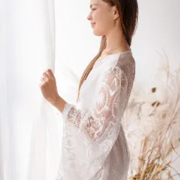 Kobieta w koronkowej sukience patrzy przez okno, profil, delikatne oświetlenie, dekoracyjne trawy w tle.