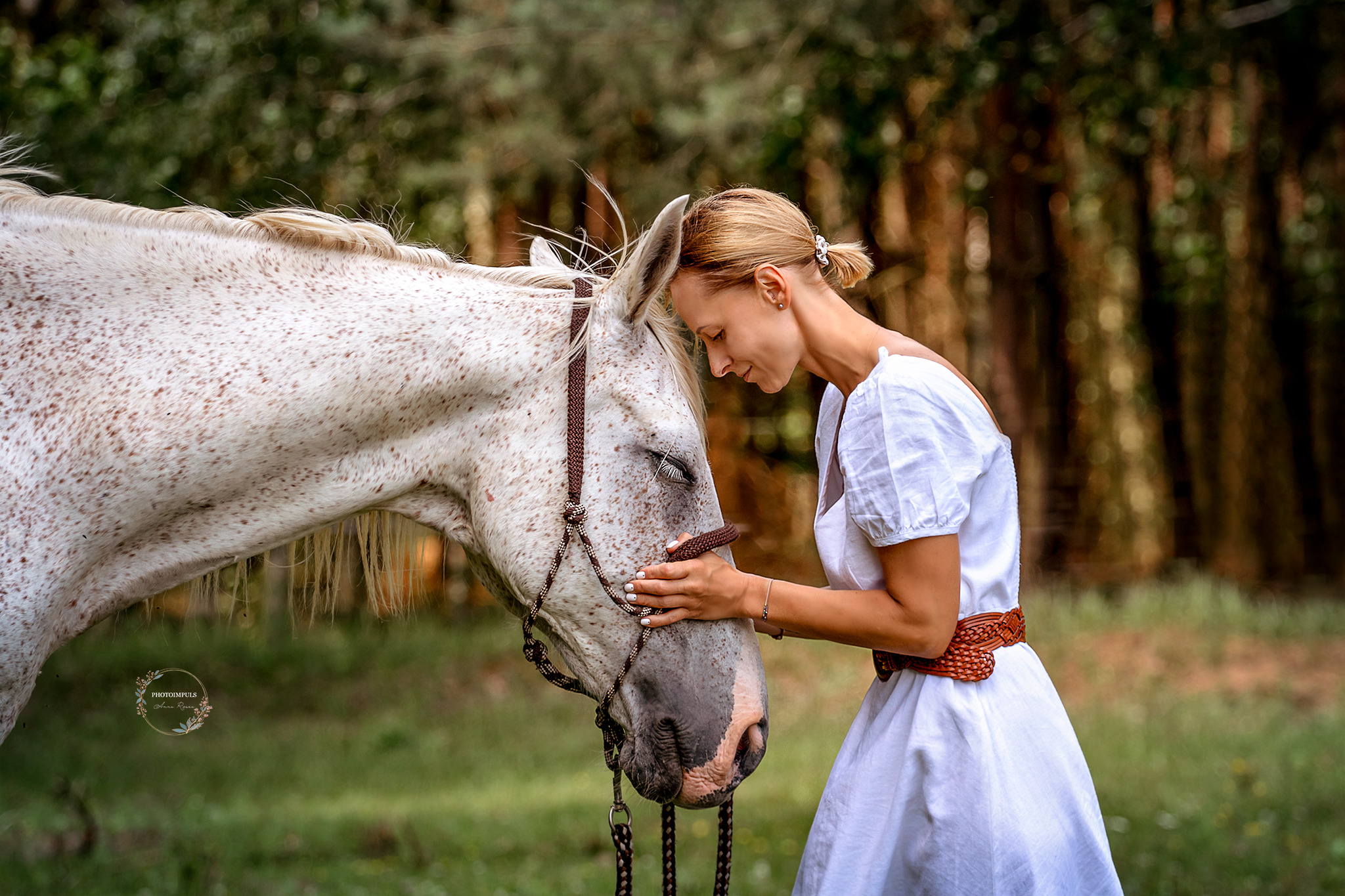 Kobieta w białej sukience, z plecionym paskiem, czule dotyka pyska siwego konia w brązowym ogłowiu na tle leśnej zieleni.
