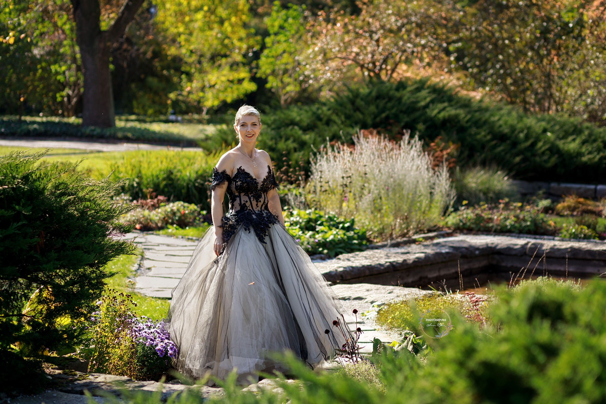 Kobieta w balowej sukni z czarną koronką pozuje w Palędziu w malowniczym ogrodzie z kamiennym obramowaniem rabat i różnorodną roślinnością.