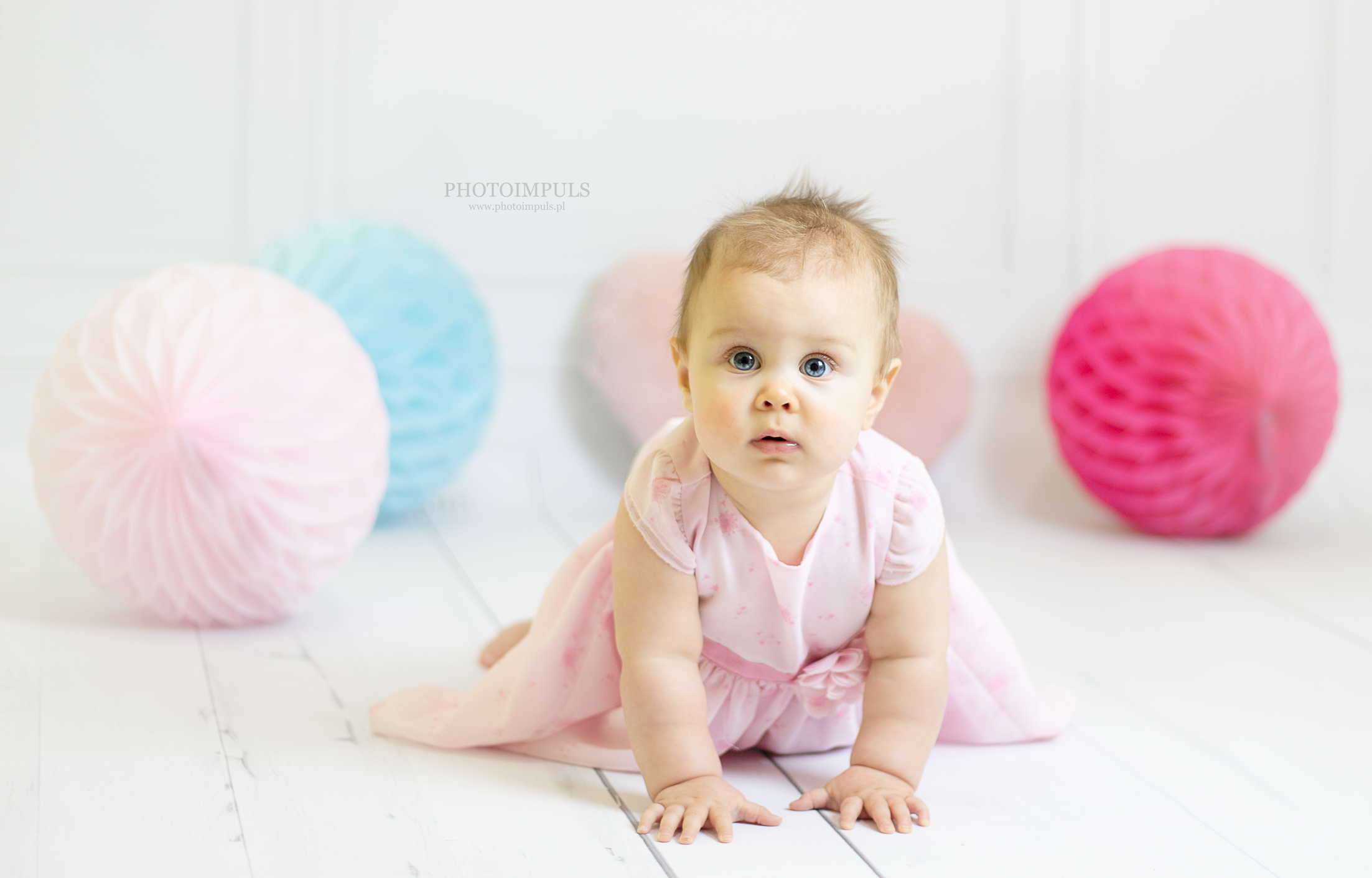 Niemowlę w różowej sukience, leżące na brzuchu na białym drewnianym podłożu, otoczone kolorowymi papierowymi kulami, patrzy prosto w obiektyw z wyrazem zaciekawienia.