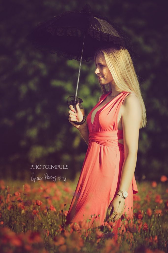 Kobieta w koralowej sukience pozuje z czarnym parasolem na łące maków w słoneczny dzień.