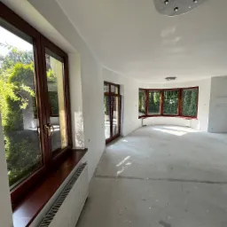 Jasne wnętrze po szpachlowaniu ścian, z drewnianymi oknami i grzejnikiem. Widoczne ślady po pracach wykończeniowych na podłodze i drabina malarska.