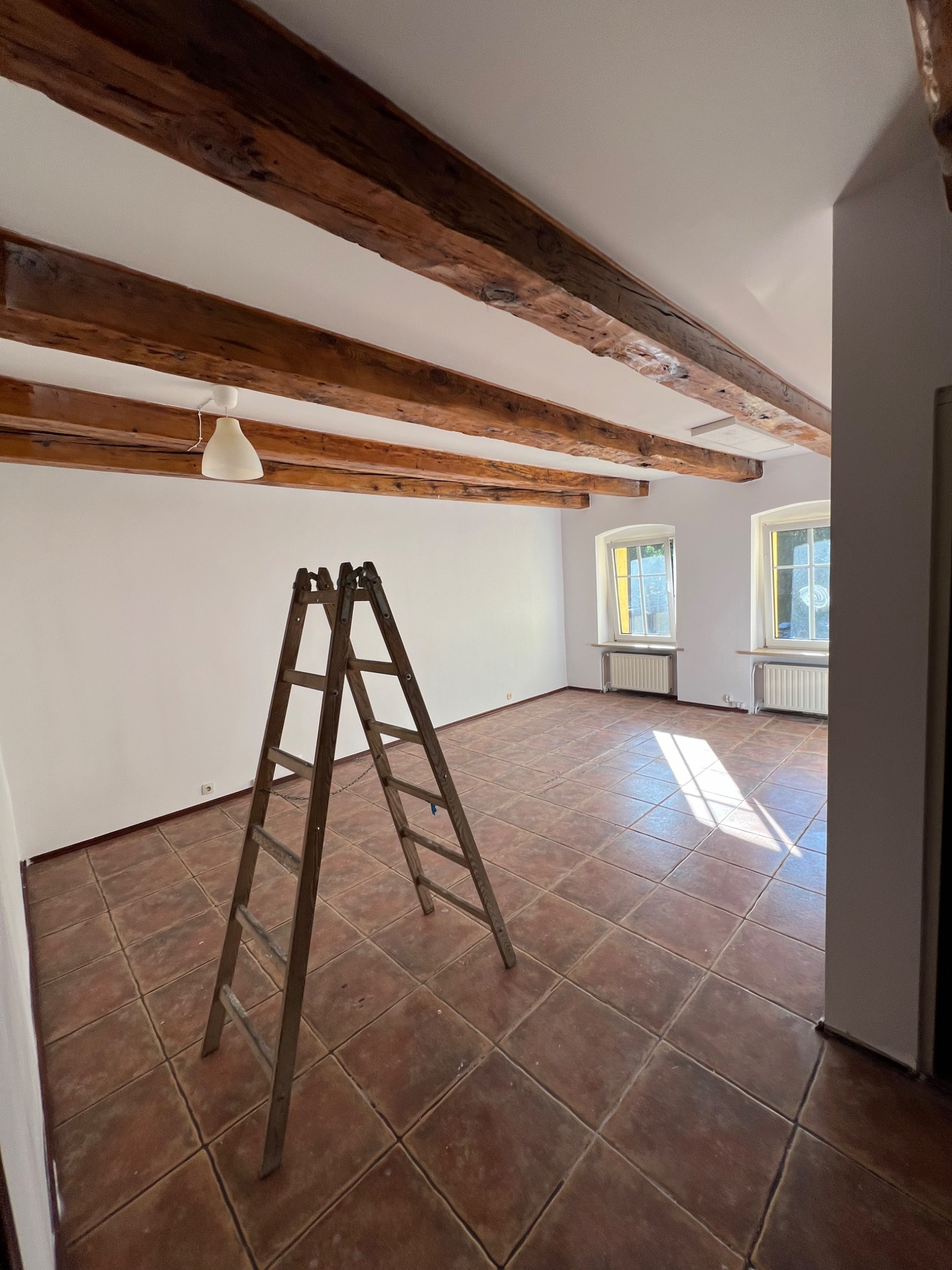 Jasne wnętrze z drewnianymi belkami stropowymi i drabiną. Ściany pomalowane na biało, terakota na podłodze. Przygotowanie do prac wykończeniowych.