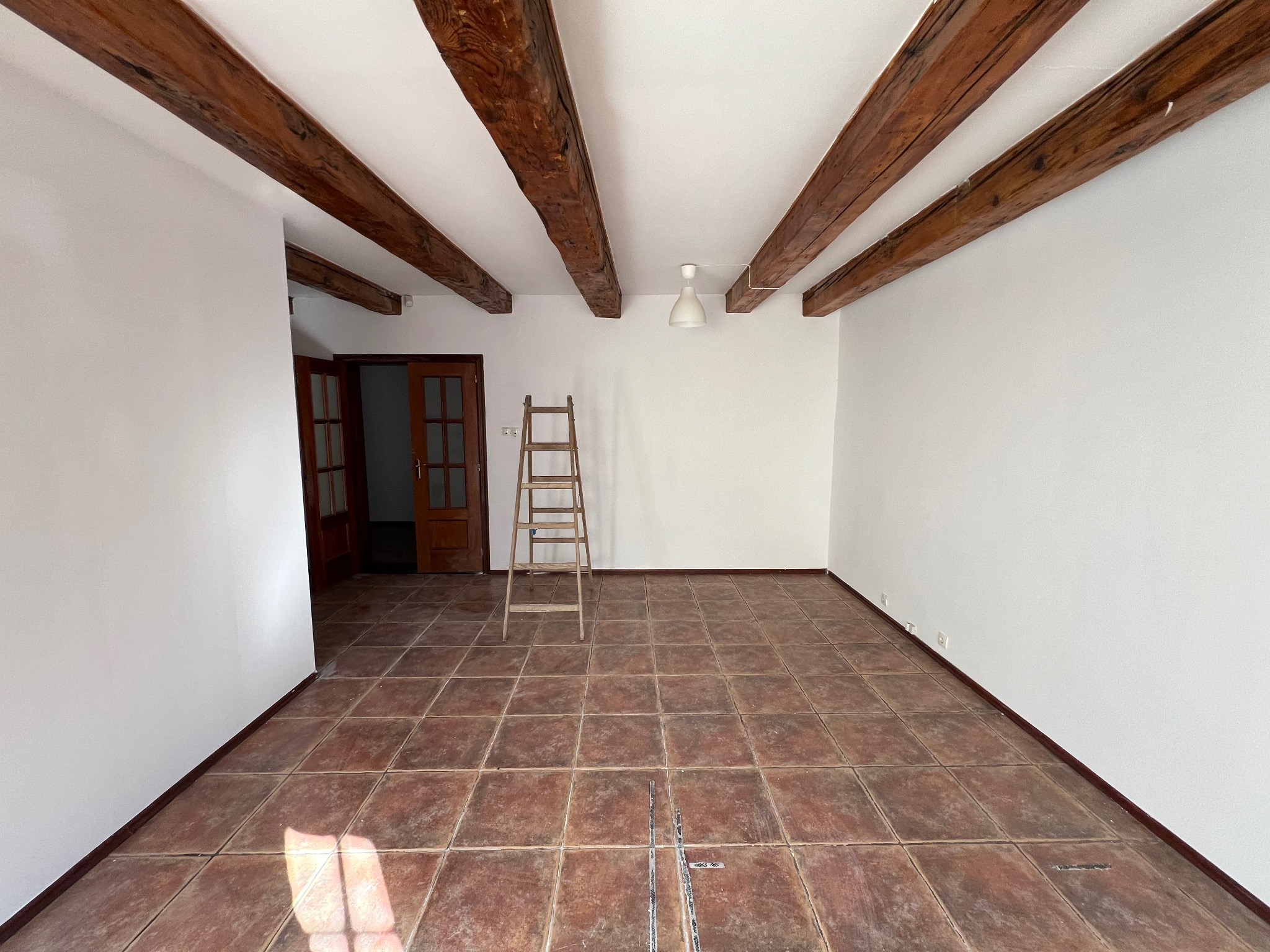 Jasne wnętrze z drewnianymi belkami stropowymi i terakotą. Drabina oparta o ścianę, przygotowania do prac wykończeniowych w przestronnym pomieszczeniu.