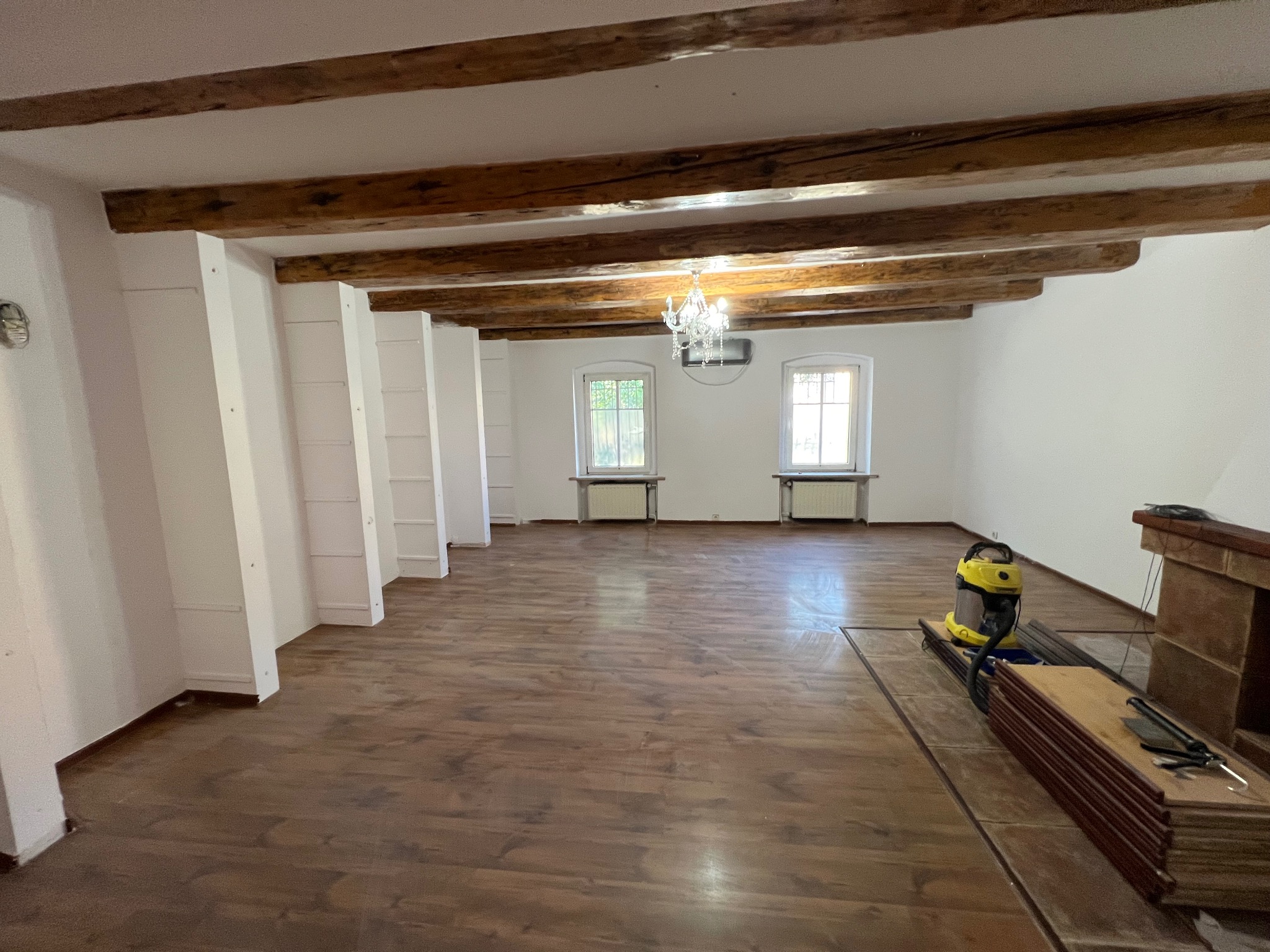 Przestronne wnętrze z drewnianymi belkami stropowymi, nową podłogą, odkurzaczem i materiałami wykończeniowymi. Widok na okna i żyrandol.
