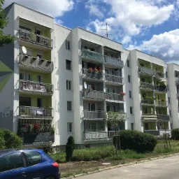 Wielorodzinny budynek mieszkalny po renowacji, z balkonami ozdobionymi kwiatami i zielenią, widoczne anteny satelitarne, elewacja w odcieniach bieli i zieleni, błękitne auto na pierwszym planie.