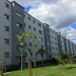 Długi blok mieszkalny po odświeżeniu elewacji w odcieniach szarości, z młodymi drzewkami na pierwszym planie i błękitnym niebem z chmurami w tle.