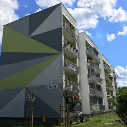 Wielopiętrowy budynek mieszkalny z odnowioną elewacją w geometryczne wzory w odcieniach szarości i zieleni, balkony z kwiatami i markizami, błękitne niebo z chmurami w tle.