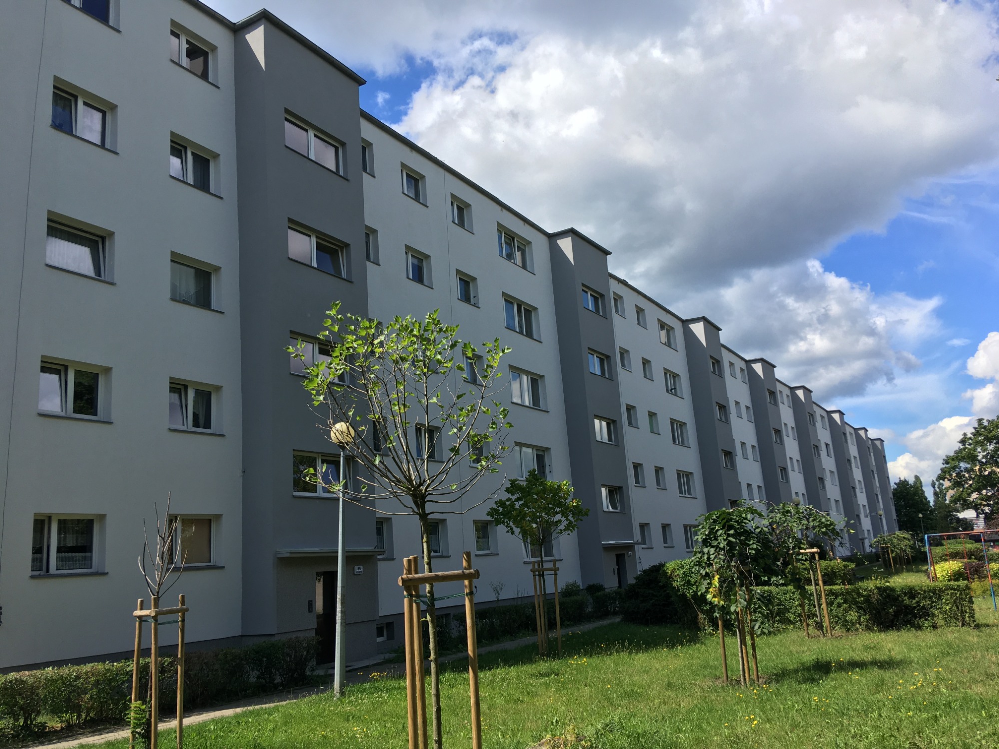 Długi blok mieszkalny po odświeżeniu elewacji w odcieniach szarości, z młodymi drzewkami na pierwszym planie i błękitnym niebem z chmurami w tle.