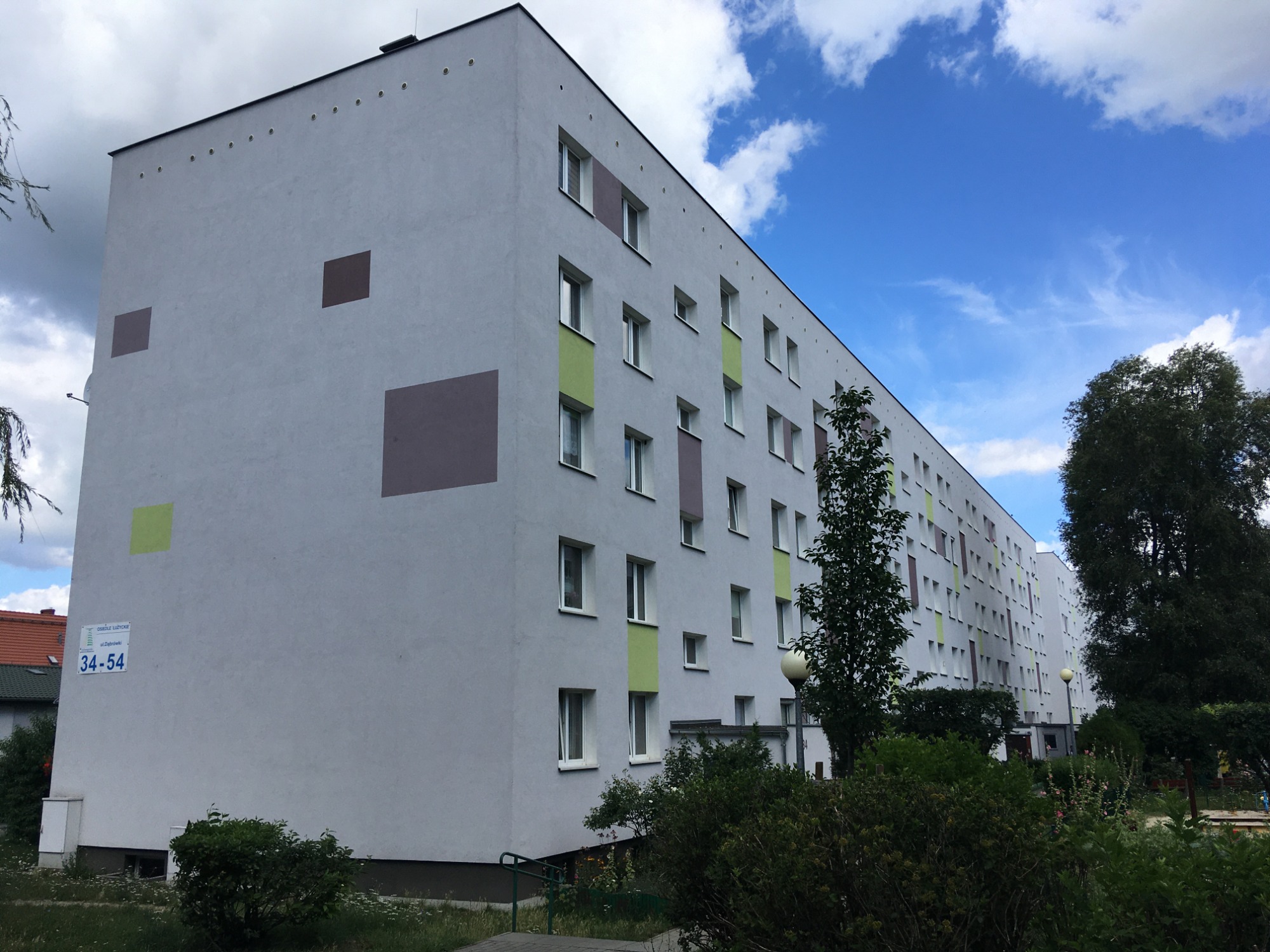 Wielopiętrowy blok mieszkalny z szarą elewacją, oznaczony kolorowymi kwadratami i ramami okiennymi w odcieniach zieleni i brązu, widoczny z poziomu trawnika w słoneczny dzień.