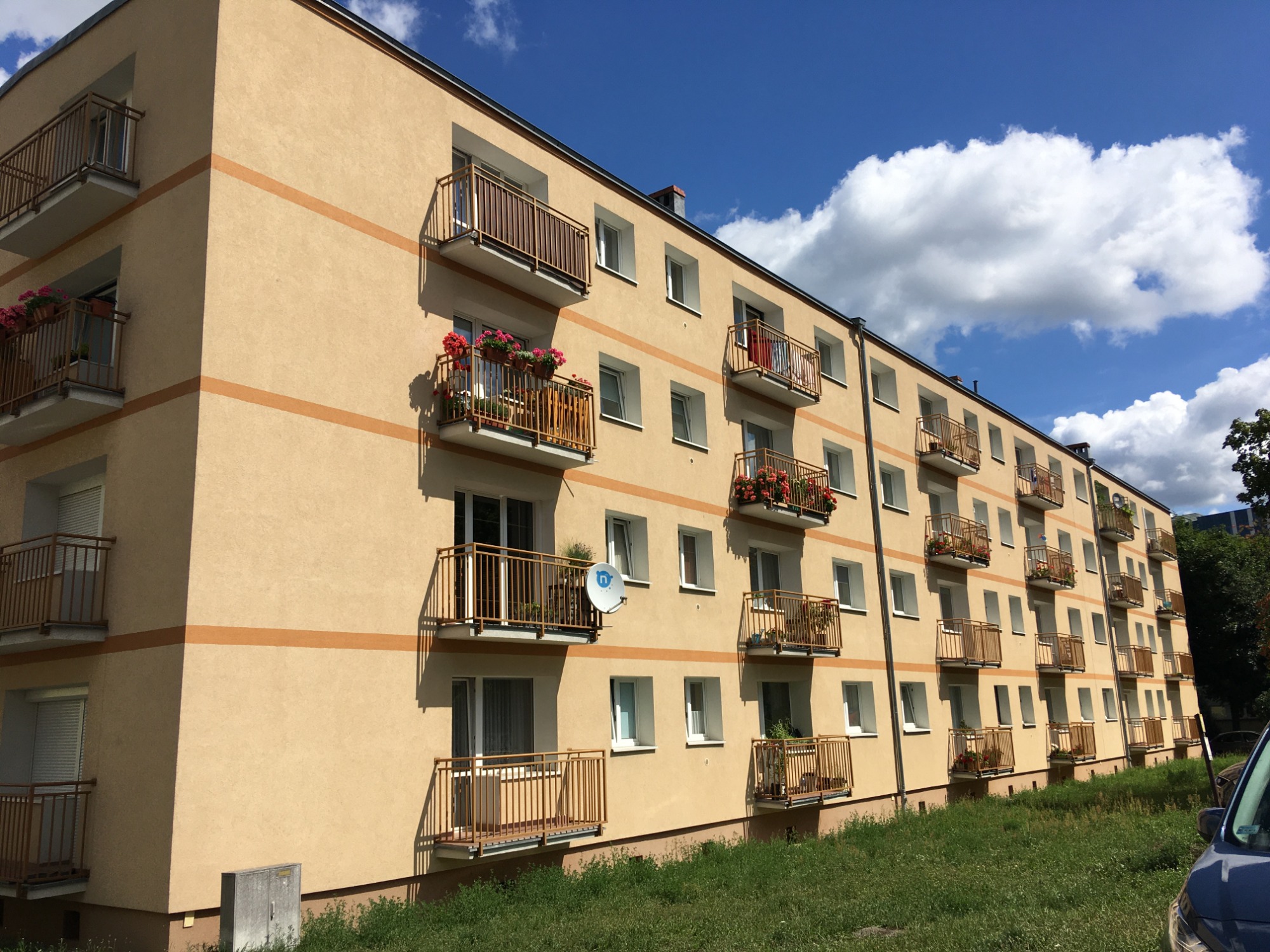 Wielopiętrowy budynek z beżową elewacją, brązowymi balkonami z kwiatami i widoczną anteną satelitarną, na tle błękitnego nieba z białymi chmurami.