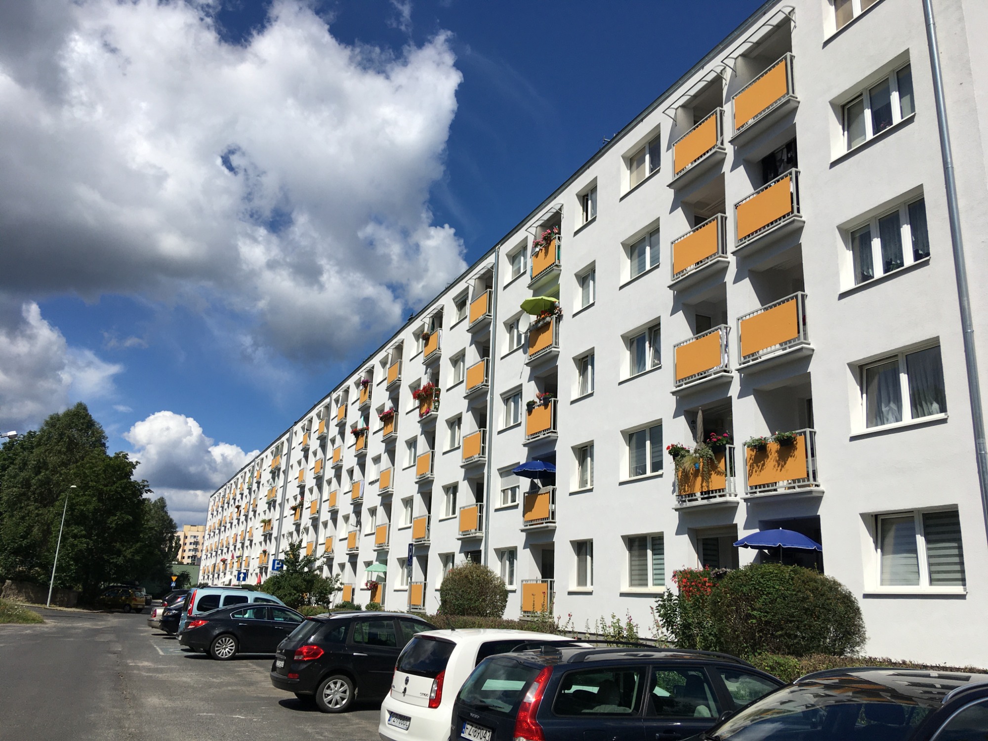 Długi, biały blok mieszkalny z rzędami pomarańczowych balkonów, niektóre z kwiatami i parasolami, na tle błękitnego nieba z obłokami, z zaparkowanymi samochodami wzdłuż ulicy.