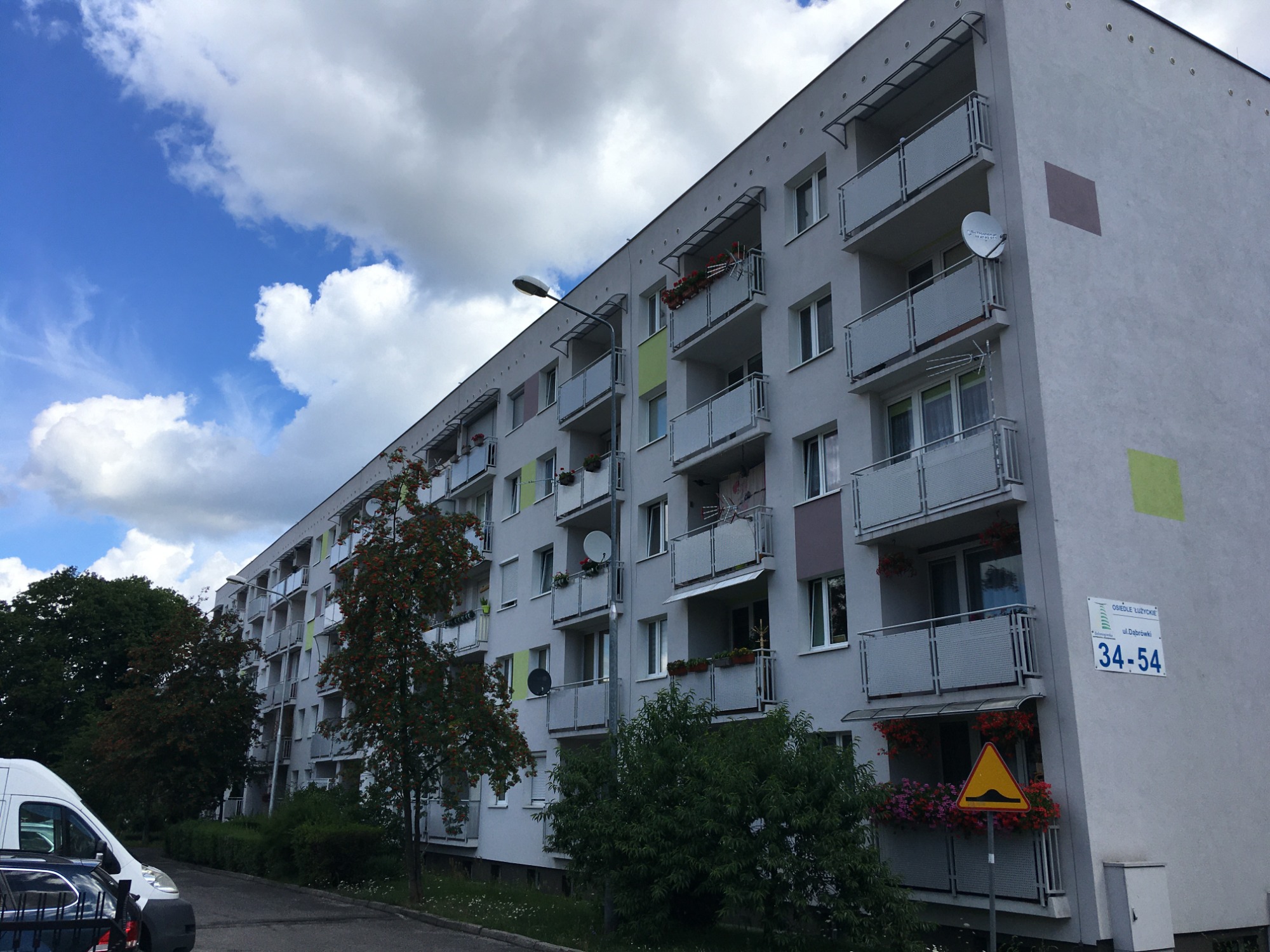 Długi blok mieszkalny z balkonami, częściowo odnowioną elewacją z kolorowymi akcentami, antenami satelitarnymi i kwiatami na balkonach, widziany z perspektywy ulicy w pochmurny dzień.