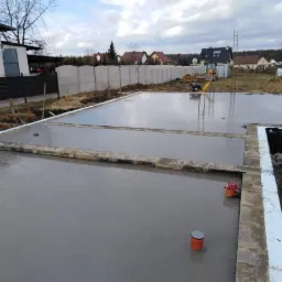 Wylany fundament z izolacją pionową, widoczne szalunki i studzienki kanalizacyjne, w tle teren budowy z koparką.