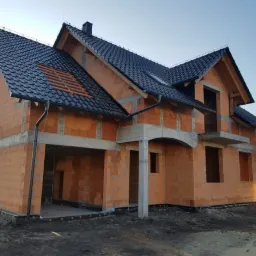 Dwukondygnacyjny dom w stanie surowym otwartym z czerwonej cegły, z czarną dachówką i widocznymi elementami betonowymi, osadzony na przygotowanym terenie.