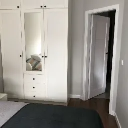 Biała szafa z lustrem wbudowanym w środkowe drzwi, widoczna w sypialni z szarą pościelą i otwartymi białymi drzwiami do innego pomieszczenia.