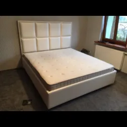 Białe łóżko tapicerowane z wysokim, kwadratowym zagłówkiem w pokoju z szarą podłogą i oknem.