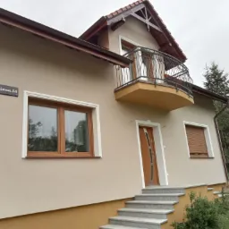 Beżowy dom jednorodzinny z balkonem z ozdobną, kutą balustradą, brązowymi oknami i drzwiami, widoczny adres na ścianie.