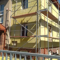 Dom w trakcie prac, widoczne rusztowanie, ocieplenie z żółtej wełny mineralnej na części elewacji, zabezpieczenie podłoża żółtą folią, fragment ceglanej ściany, niebieskie niebo w tle.