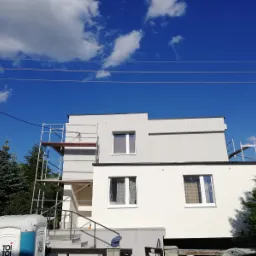 Dwukondygnacyjny dom w trakcie prac elewacyjnych: część ściany pomalowana na szaro, reszta na biało, widoczne rusztowania i przenośna toaleta na pierwszym planie.