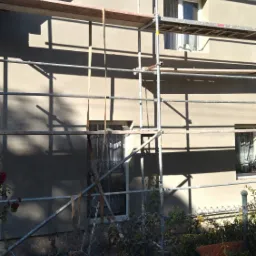 Dom jednorodzinny w trakcie prac elewacyjnych, widoczne rusztowanie, świeżo nałożony tynk w kolorze beżowym, okna zabezpieczone folią, ogród z przodu budynku.