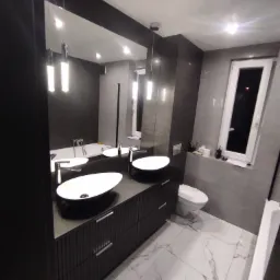 Nowoczesna łazienka z dwoma owalnymi umywalkami nablatowymi, czarną szafką z frezowanymi frontami, lustrem sięgającym sufitu, szarymi płytkami na ścianach i podłogą z białego marmuru z szarymi żyłami.