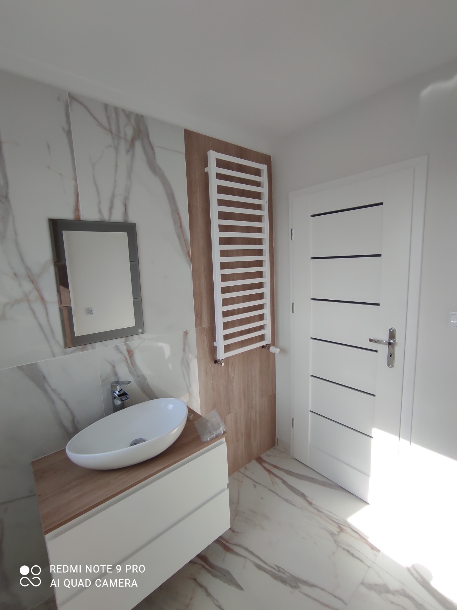 Nowoczesna łazienka z owalną umywalką nablatową, lustrem w grafitowej ramie, białym grzejnikiem drabinkowym i białą, minimalistyczną szafką pod umywalką na tle płytek imitujących marmur.