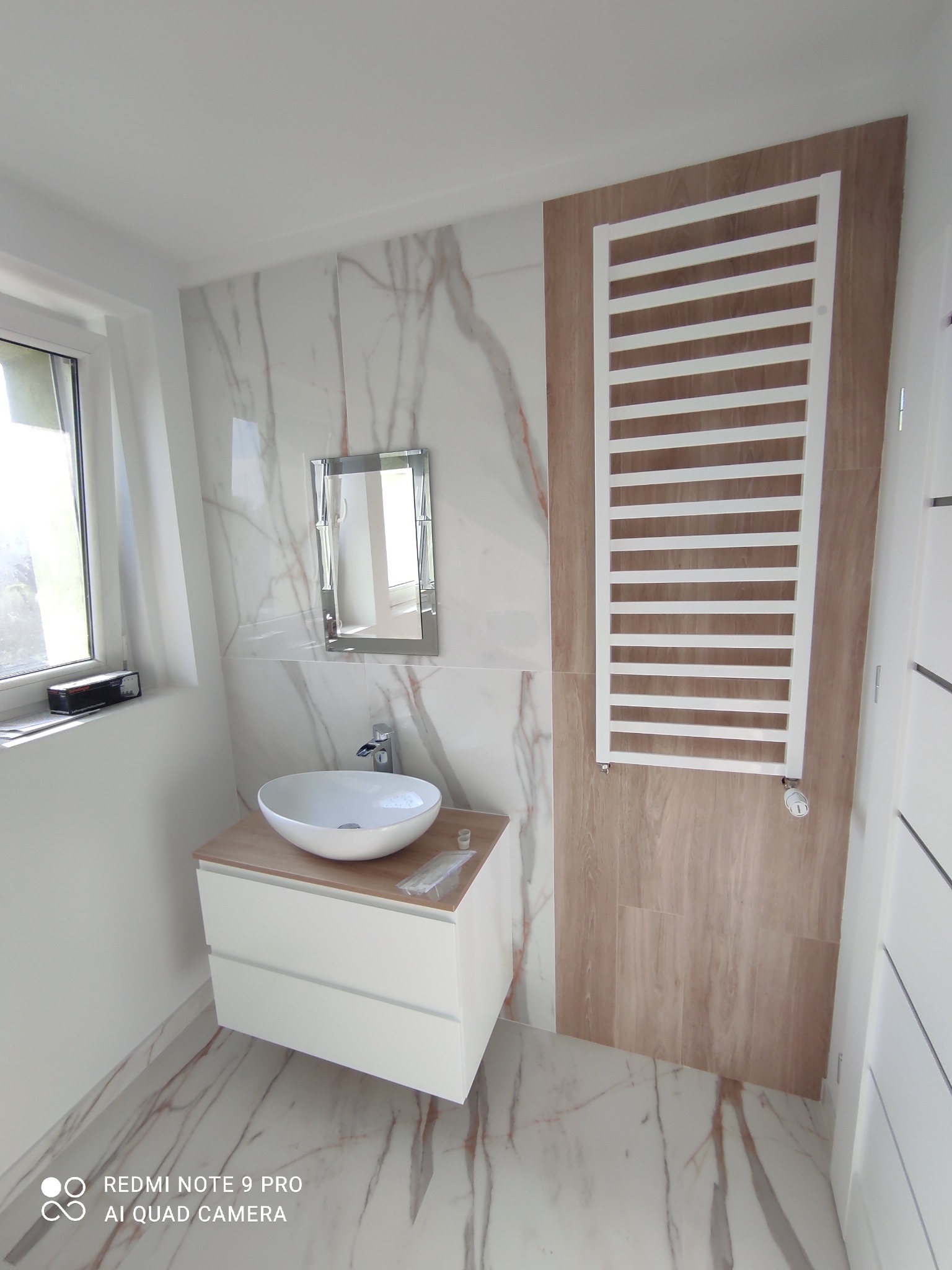 Nowoczesna łazienka po remoncie: marmurowe płytki na ścianach i podłodze, biała szafka z umywalką nablatową, grzejnik drabinkowy na tle drewnopodobnych paneli, lustro w cienkiej ramie, widok z góry.