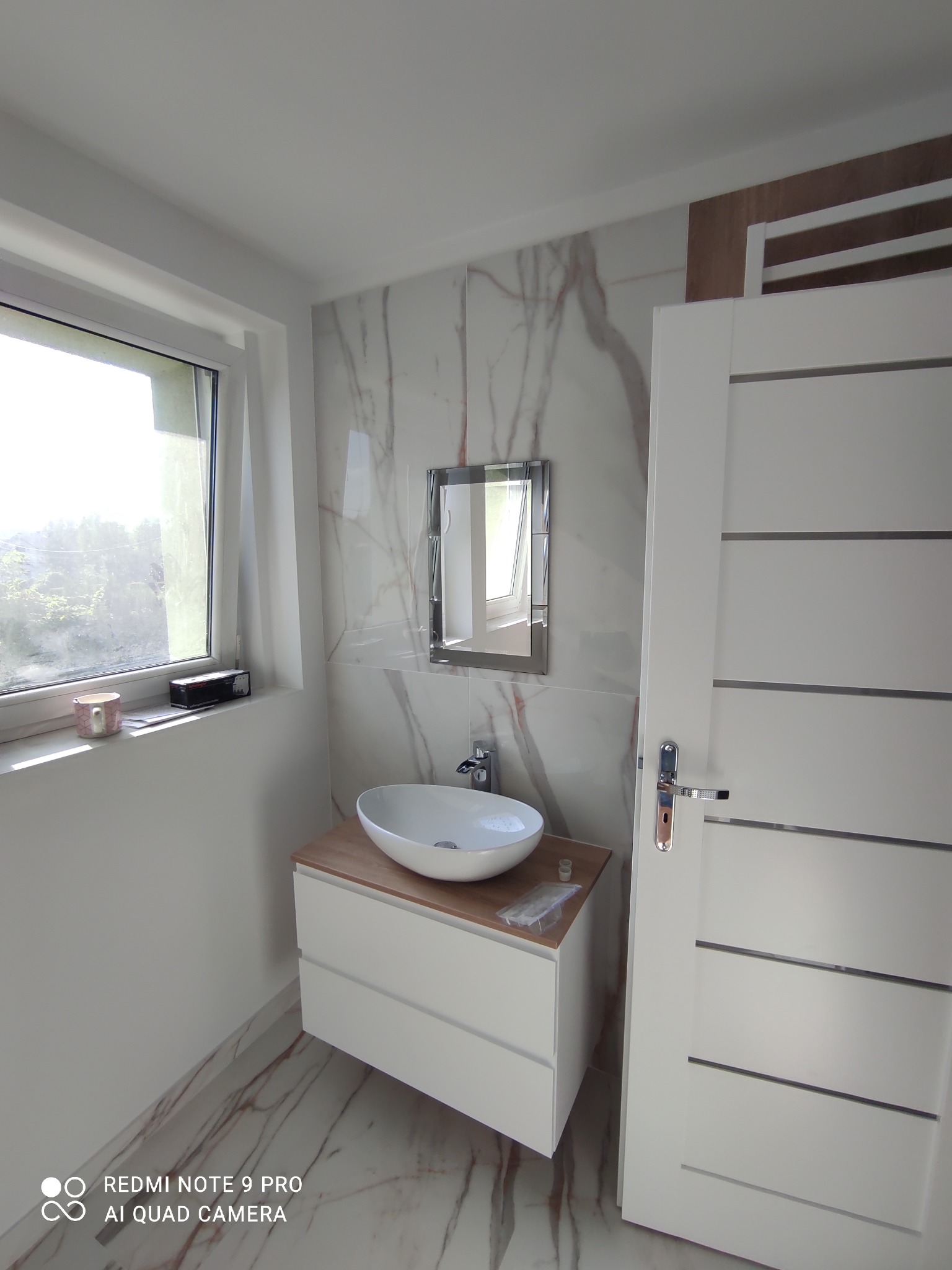 Nowoczesna łazienka z marmurowymi płytkami, owalną umywalką nablatową na szafce z drewnianym blatem, lustrem w ramie i białymi drzwiami z poziomymi wstawkami.