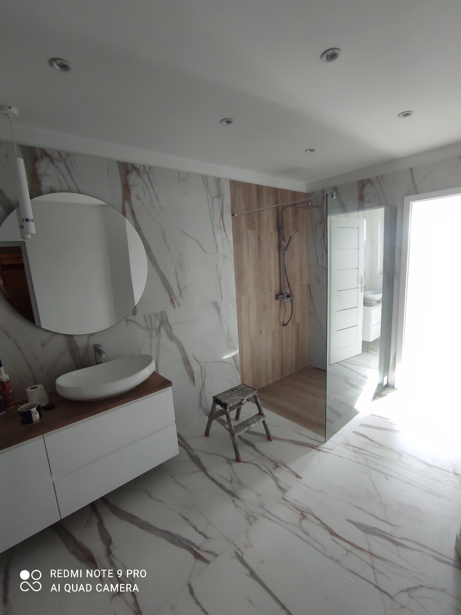 Wykończona łazienka z marmurowymi płytkami, okrągłym lustrem nad białą szafką z umywalką, szklanym prysznicem i drewnianym akcentem na ścianie, widoczny stołek malarski.