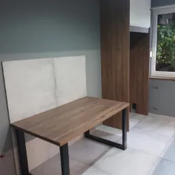 Stół z drewnianym blatem i czarnymi nogami w surowym wnętrzu, widoczna zabudowa szafkowa z elementami drewnopodobnymi i białymi frontami, szara ściana i płytki podłogowe.