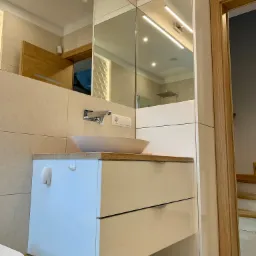 Nowoczesna łazienka z umywalką nablatową w odcieniach beżu i bieli, z drewnianym blatem i lustrem z oświetleniem LED, widoczne schody w tle.