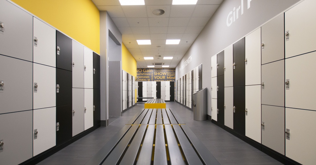 Długi korytarz z rzędami szafek w szatni, utrzymanych w odcieniach szarości, bieli i czerni, z żółtym akcentem na ścianie i ławkach, oraz napisem motywacyjnym na końcu korytarza.