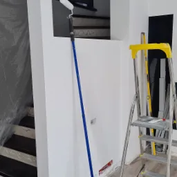 Świeżo pomalowane na biało ściany klatki schodowej z widocznym wałkiem malarskim opartym o ścianę, drabina aluminiowa z żółtymi elementami stoi obok, zabezpieczone schody w tle.