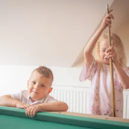 Dwoje dzieci: chłopiec oparty o stół bilardowy, uśmiechnięty, i dziewczynka z blond włosami trzymająca kij bilardowy pionowo nad stołem.