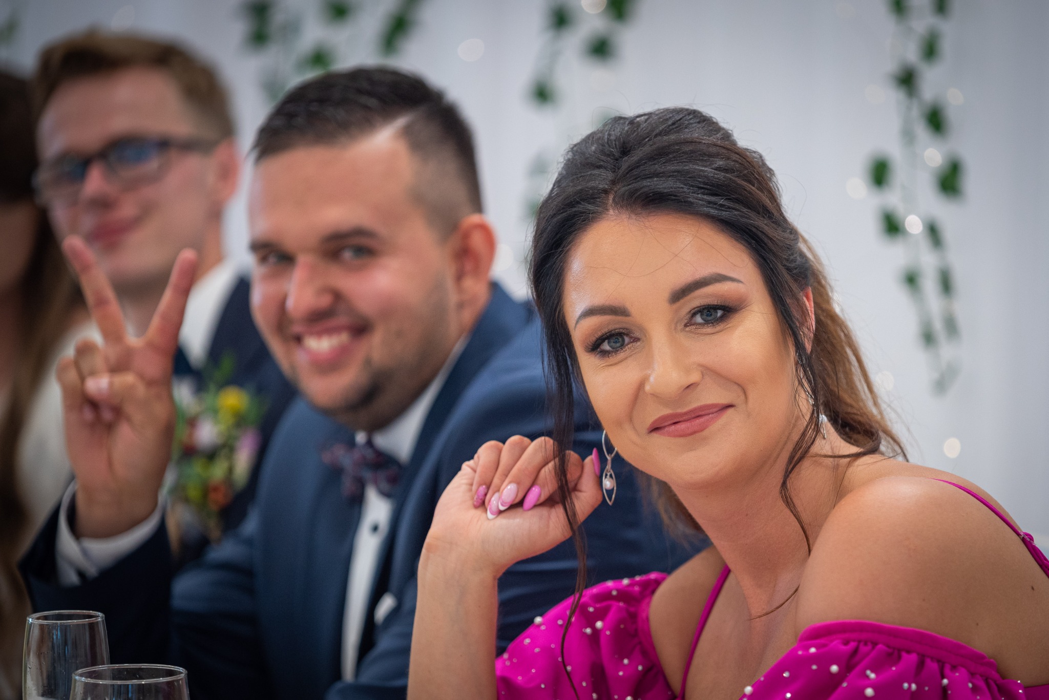 Uśmiechnięta panna młoda w fuksjowej sukience z perłami, obok pan młody w granatowym garniturze z muszką i gość z symbolem zwycięstwa na weselu, w tle dekoracje świetlne.